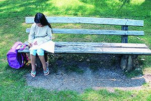 Ein Mädchen sitzt alleine auf einer Parkbank und erledigt seine Hausaufgaben. Neben der Bank steht der Schulranzen des Mädchens.